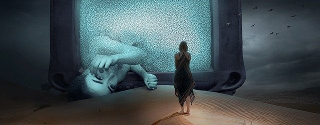 huge screen sitting in a desert showing a sad girl laying on her side and a girl standing in front of the screen watching